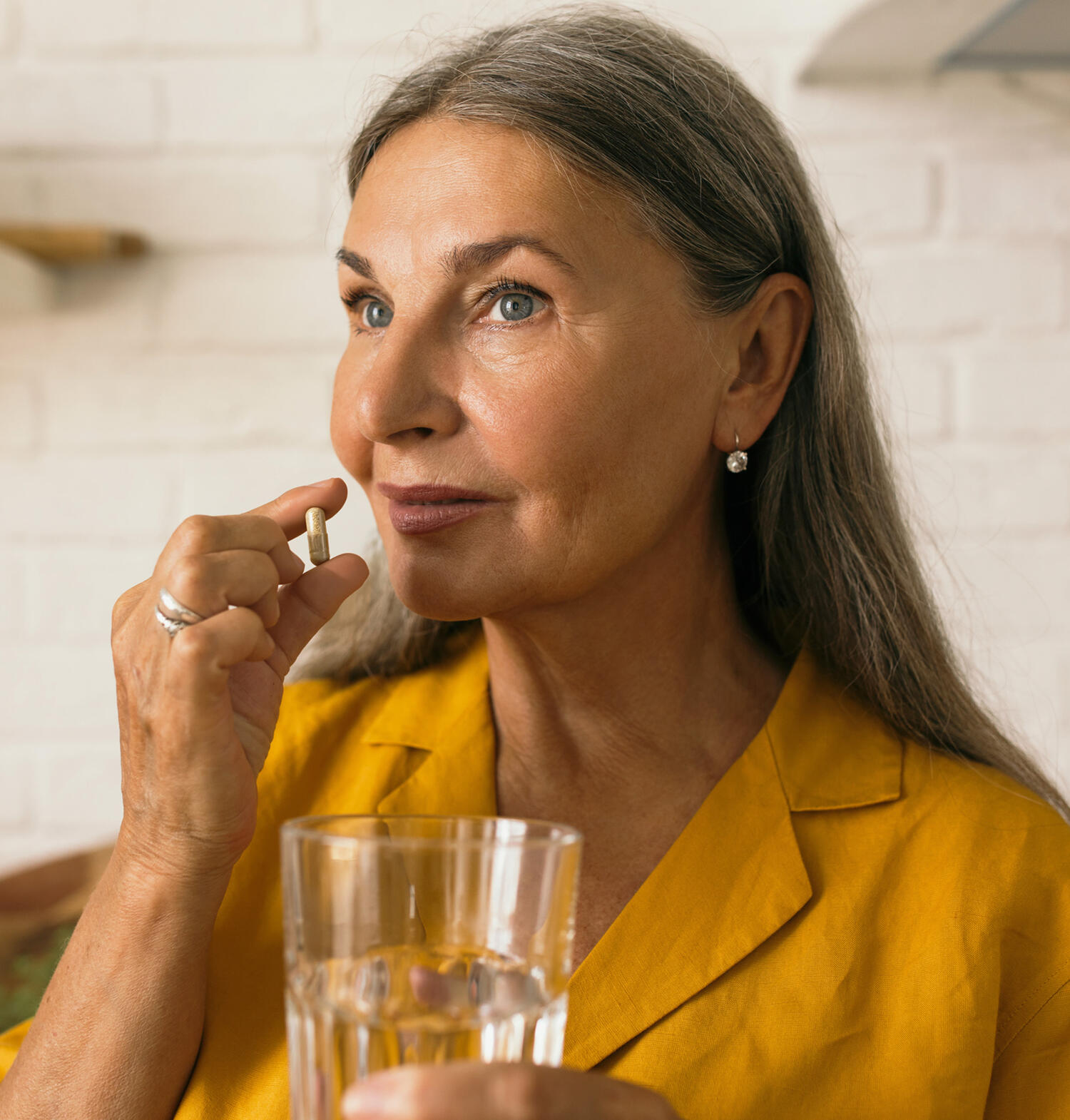 Older woman with gray eyes and long gray/blonde hair wears a golden yellow button up while holding a brown supplement capsule and a glass of water