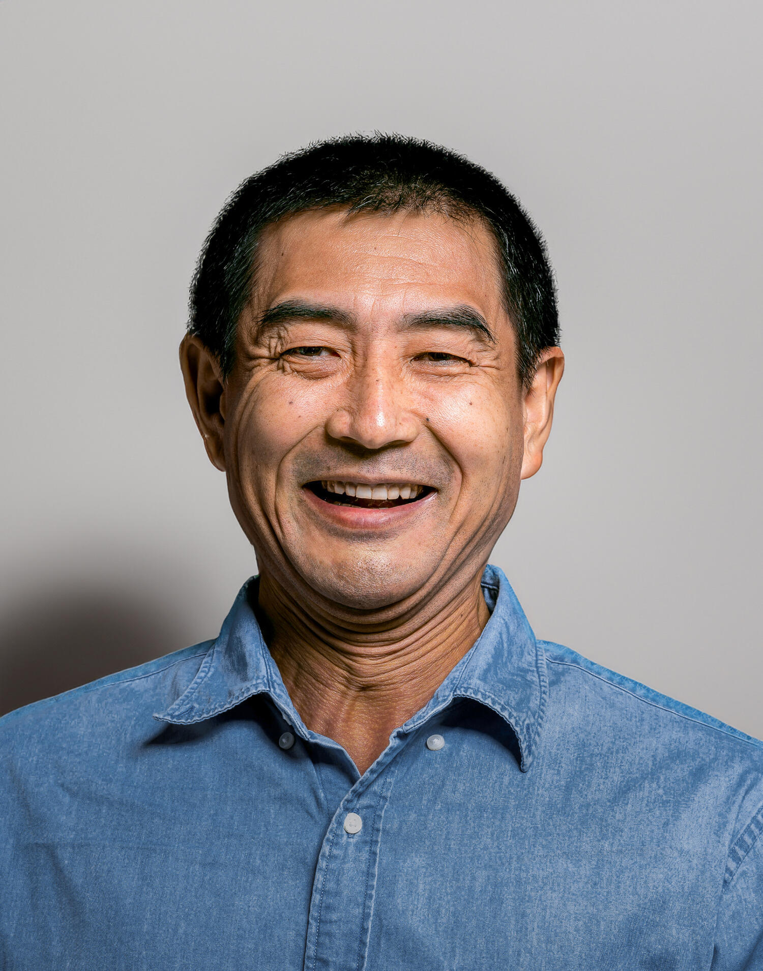 Photo of a smiling middle aged Asian man with short hair, wearing a denim button up